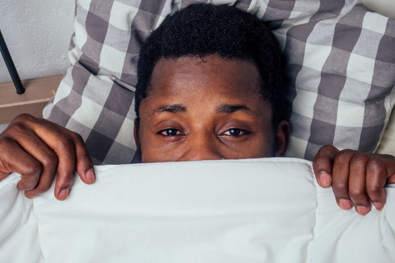 Man hiding under duvet in bed showing eyes