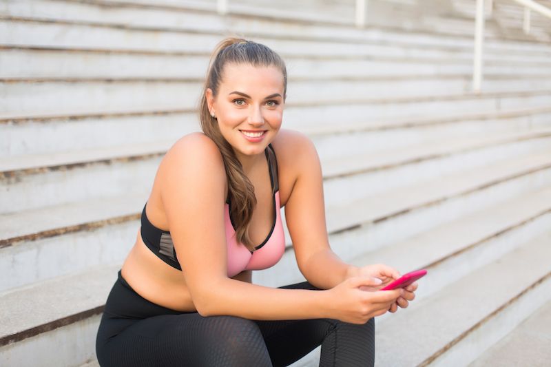 Femme en tenue de sport assise dans les escaliers et tenant son téléphone