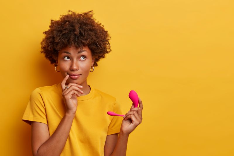 Thoughtful woman holding G-spot toy on yellow background