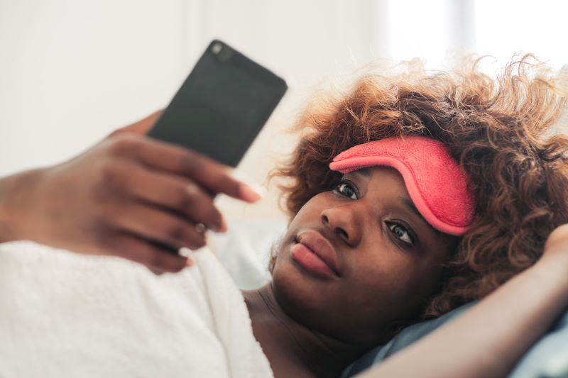 Femme avec masque pour les yeux vérifiant son téléphone au lit